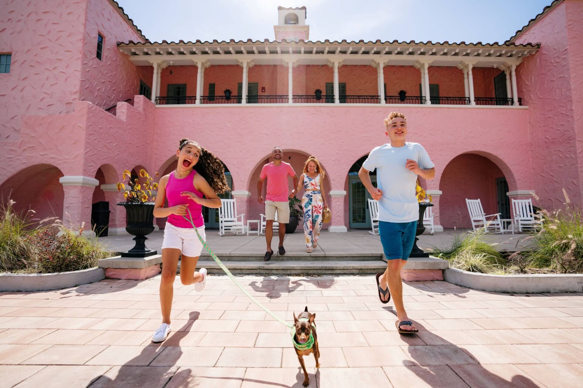 Una familia de cuatro personas sale corriendo del histórico Hotel Hacienda en el centro de New Port Richey. La familia viste ropa de diferentes colores que contrastan con el rosa brillante del hotel que tienen detrás. Los padres están al fondo, mientras que sus dos hijos van delante; su hija lleva una correa verde y su chihuahua ríe mientras salen del hotel. 